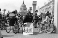 Protesters outside of the Federal Building with SF City Hall in the background. A prominent sign on the back of a long-haired man's wheelchair reads "We shall overcome."